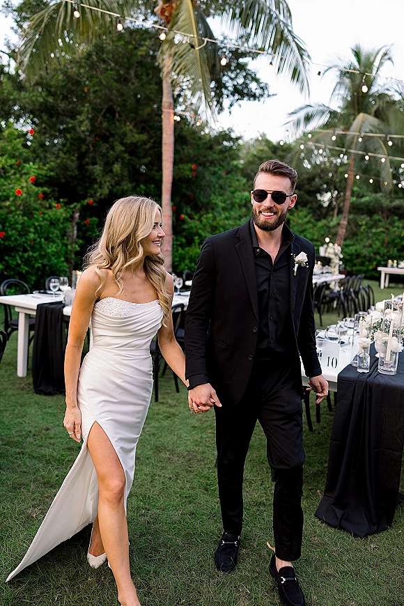 Couple portrait of newlyweds holding hands, bride in strapless satin slit gown and groom in black suit under string lights by palms