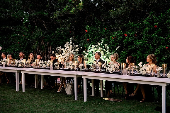 Wedding head table with outdoor head table decor, long banquet table of white florals, greenery, and glass cylinder candles in a garden setting