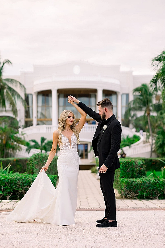 Couple portrait of bride twirling as groom leads her spin, lace dress train flowing on stone walkway with palm trees and grand facade.