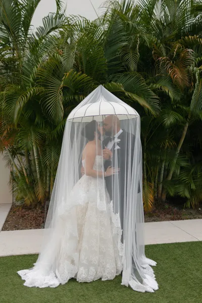 Couple portrait of bride and groom under veil sharing an almost-kiss, his white boutonniere prominent, with palm trees lining a grass lawn.