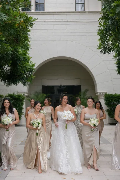 Bride with bridesmaids in champagne bridesmaid dresses, the bride holding white orchids, gathered by a stone arched doorway in a courtyard.