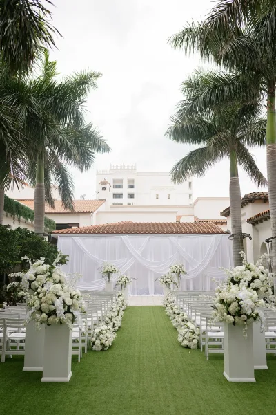 Ceremony setup with white ceremony decor, white florals on pedestal stands lining a green aisle in a palm-lined courtyard.