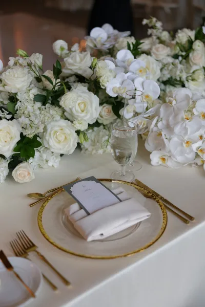 Reception tablescape with a white floral runner centerpiece and gold rim glassware under soft ambient lighting