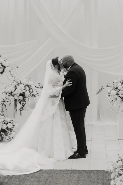 Ceremony kiss moment as a couple at the altar embraces beneath a draped fabric backdrop, bride in a lace wedding dress and train on the tile floor.