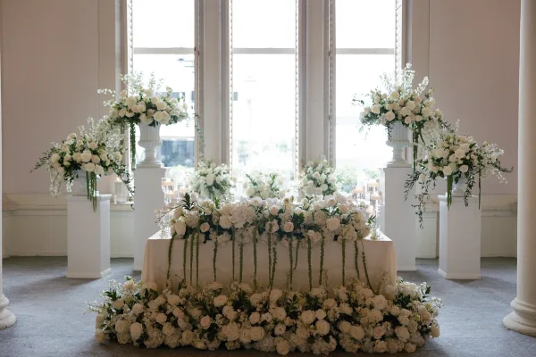 Sweetheart table decor with a white rose table garland and floating candles, set before large windows and indoor columns