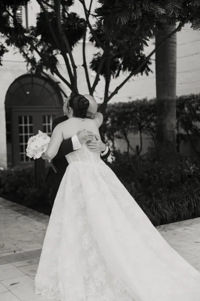 Wedding first look as the bride and groom embrace, her lace bridal gown sweeping the stone patio in a courtyard by an arched doorway.