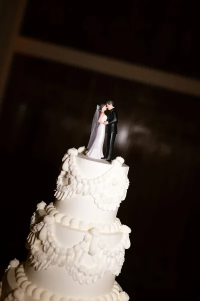 Wedding cake, a white tiered wedding cake crowned with a bride and groom cake topper, set against a dark indoor wall.