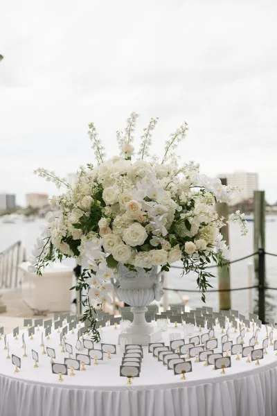 Escort card display, an elegant place card display topped with white florals, set beside a waterfront marina with docks under a cloudy sky.