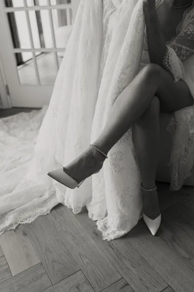 Bridal shoes, white wedding heels, rest beside a lace robe on a hardwood floor by bright French doors.