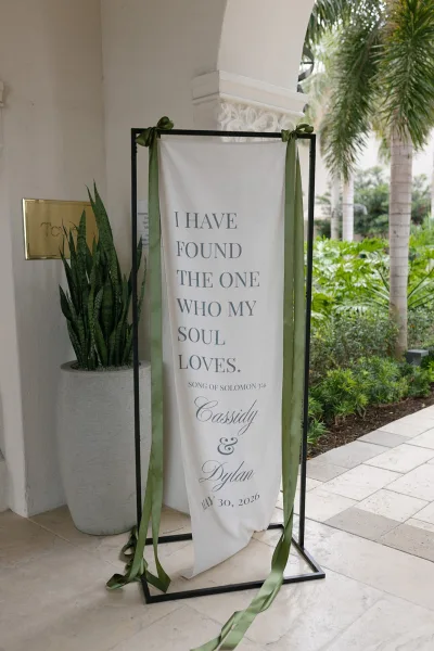Wedding sign, a fabric wedding sign on a metal frame, set under a stucco archway with palm trees and garden greenery on stone tiles.
