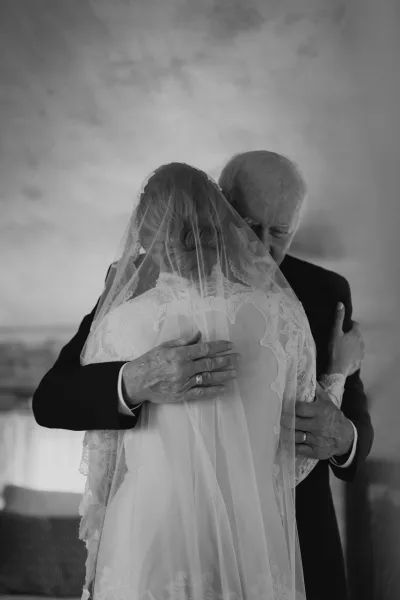 Father daughter hug during a first look, bride in lace dress and long veil embracing dad in suit by an indoor sofa