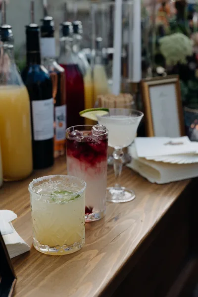 Wedding cocktail bar with signature wedding cocktails, lime-garnished glasses and dispensers beside a framed menu on a rustic wood top