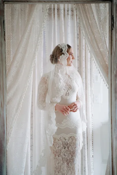 Bridal portrait of a bride by window in a lace wedding dress, long sleeves and veil, lit by soft window light through lace curtains