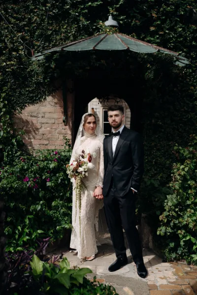 Couple portrait of bride and groom holding hands, her lace wedding dress and veil by an ivy-covered stone archway outdoors