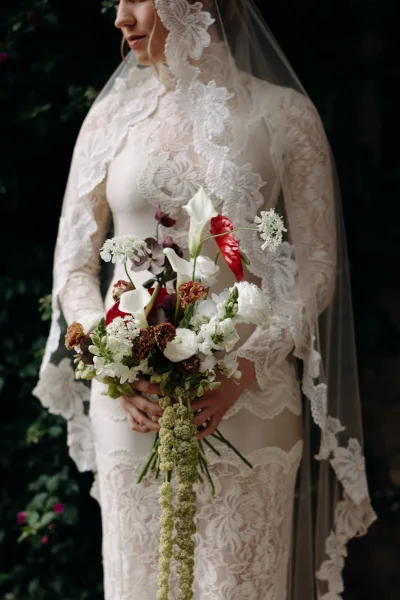 Bridal portrait of a bride holding bouquet of calla lilies, anthurium, and trailing amaranthus in a long sleeve lace dress against dark greenery