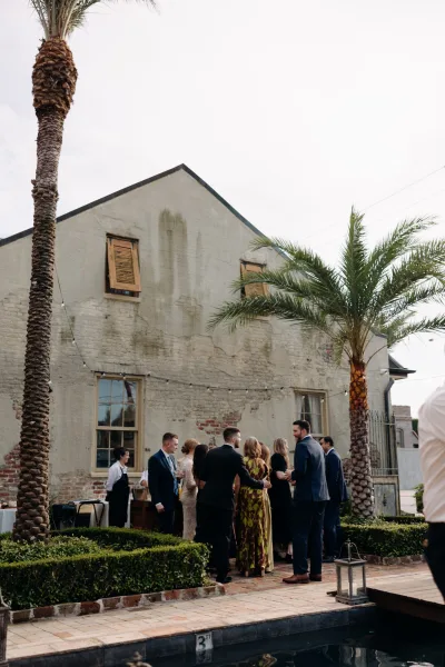 Wedding cocktail hour with wedding guests mingling in suits and a long dress, holding cocktails under string lights by a poolside brick courtyard