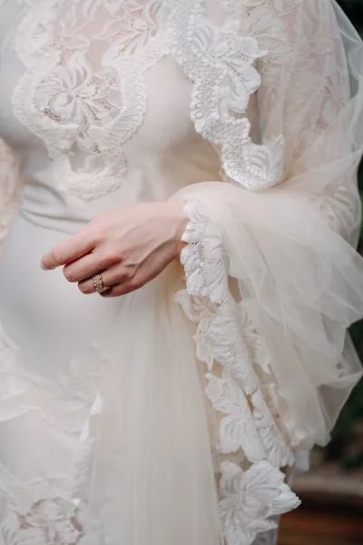 Wedding dress close-up showing lace wedding dress detail on illusion bodice with tulle sleeve and beaded ring, blurred greenery behind