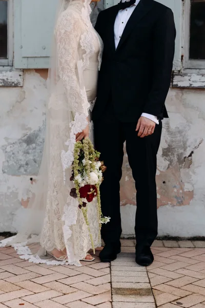 Couple portrait of bride in lace long sleeve wedding dress holding a cascading bouquet beside groom in black tuxedo against stucco wall with shutters