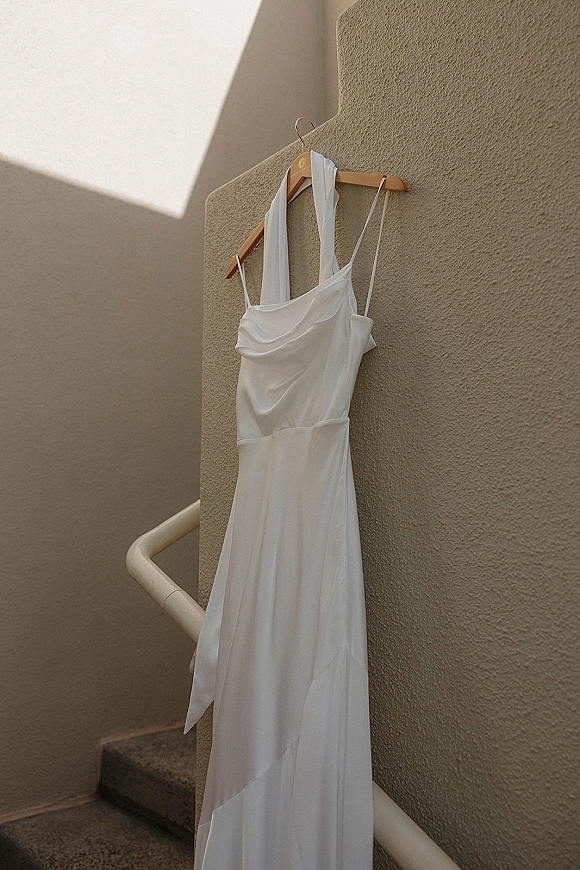 Wedding dress in satin on a wooden hanger, featuring spaghetti straps and a draped neckline, hanging by a sunlit stucco stairwell