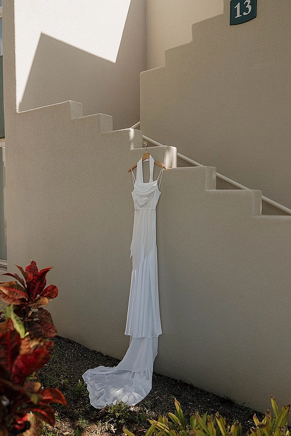 Wedding dress on hanger draped from a wooden hanger against a stucco wall by an outdoor staircase with plants and gravel ground