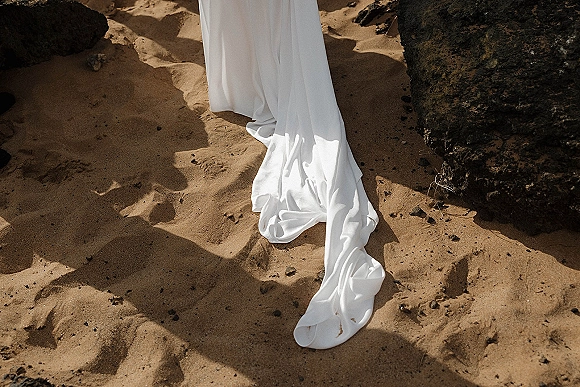 Wedding dress train of a white bridal gown trailing over sand by a rocky beach, with sharp shadows highlighting the fabric folds