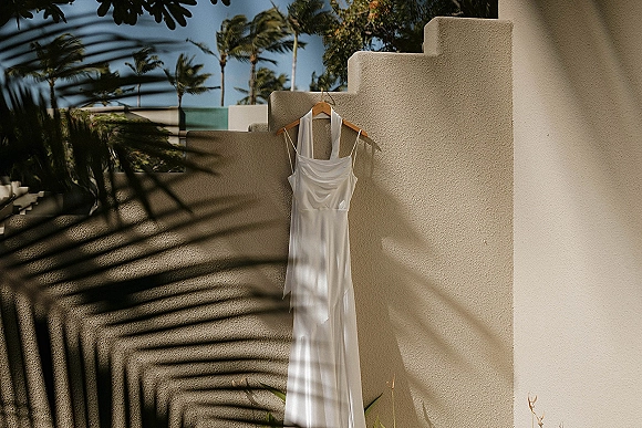 Wedding dress on a wooden hanger, a simple wedding dress in white, hanging against a stucco wall with palm trees and sky shadows
