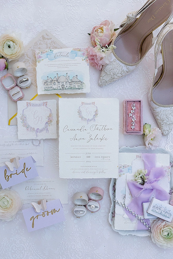 Invitation suite with watercolor wedding invitation, RSVP card, wax-seal envelope and lilac ribbon, rings, vow books on textured white surface