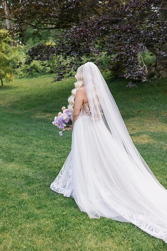 Bridal portrait of a bride from behind in a strapless lace wedding dress with long cathedral veil, holding a pastel bouquet on a garden lawn