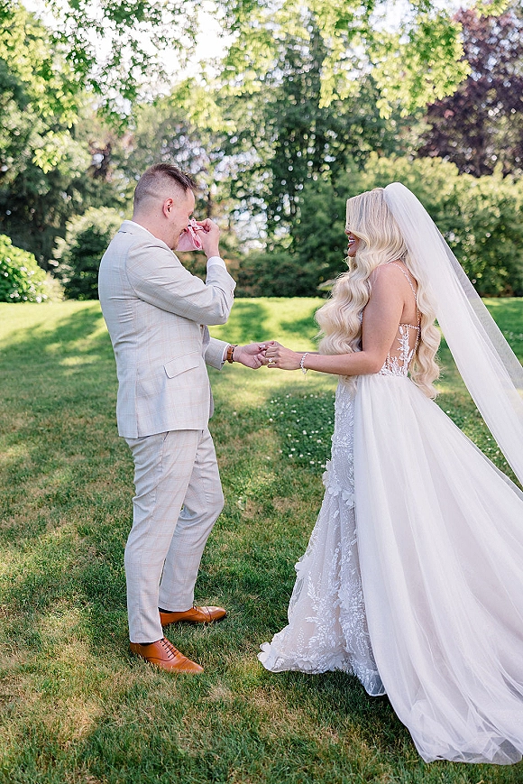 First look moment as bride in lace gown and cathedral veil reaches for groom wiping tears on a sunlit garden lawn with trees