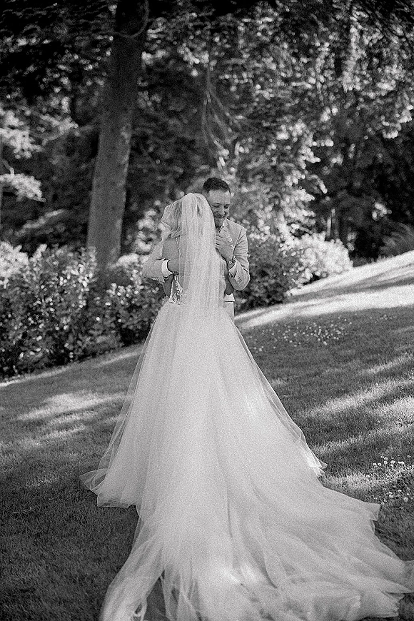 First look moment as bride in lace gown with long veil and train embraces groom in suit on a sunlit garden lawn among trees