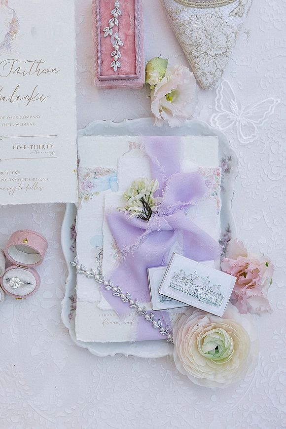 Wedding invitation suite with calligraphy wedding invitation, rings in pink box and lavender ribbon on white lace with blush blooms on tray