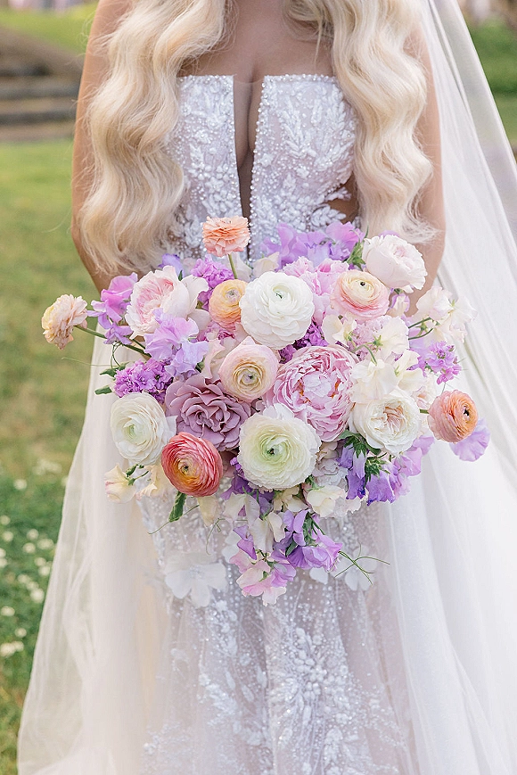 Bridal bouquet of pastel blooms, pastel wedding bouquet with peonies, ranunculus and sweet peas held against a lace dress outdoors on a lawn