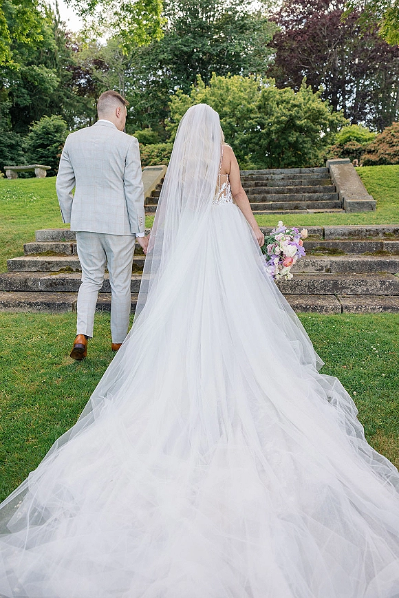 Bride and groom holding hands walk up stone steps, her cathedral veil trailing behind as she carries a pastel bouquet in a garden setting