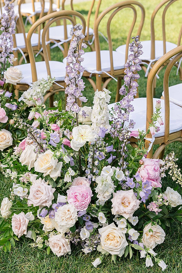 Ceremony aisle flowers with aisle meadow florals—ground roses, peonies, and greenery lining wooden chairs on a grass lawn
