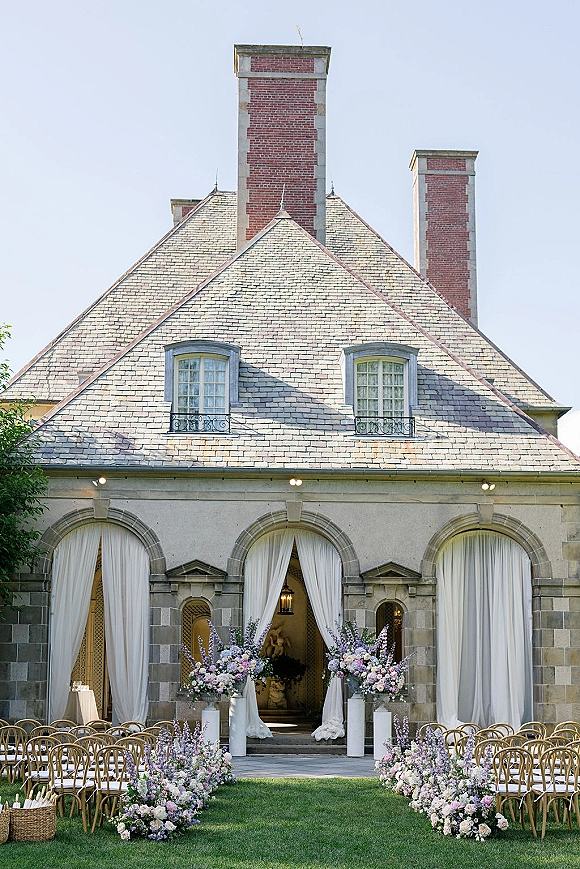 Ceremony setup with white draping and floral arrangements lining the aisle, set in a stone courtyard with arched doorways under blue sky