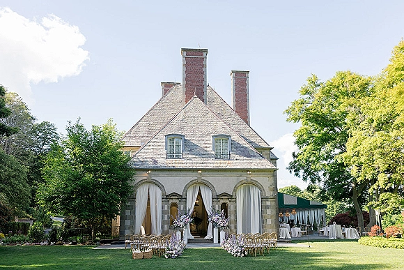 Outdoor ceremony setup with garden ceremony chairs lining a flowered aisle, facing white drapery at a stone estate under a tent canopy