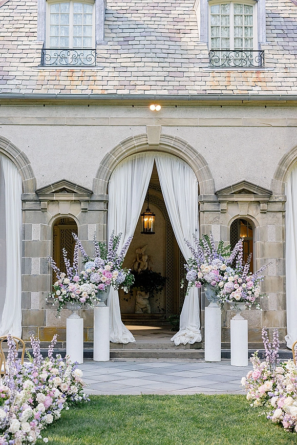 Wedding ceremony setup with outdoor ceremony backdrop, white draping and pastel floral urns framing an arched stone doorway entrance