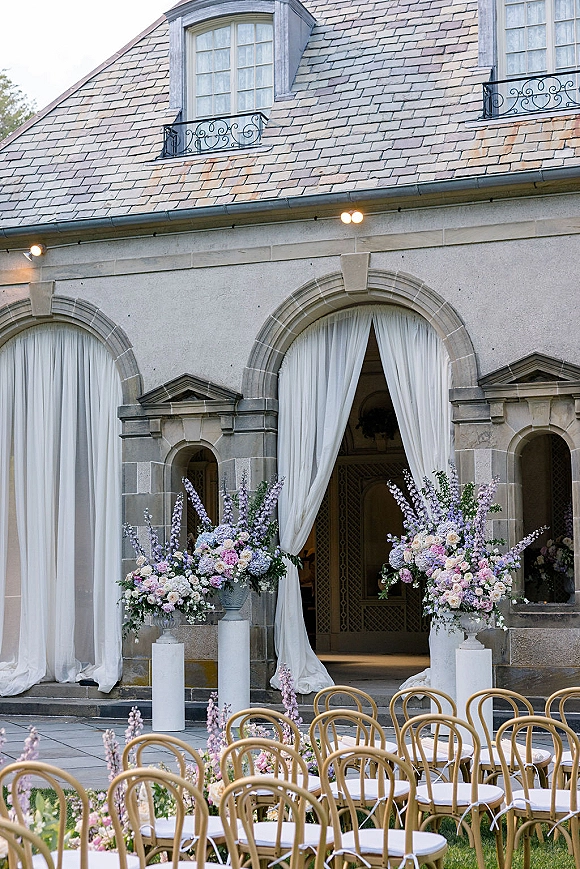 Ceremony backdrop with white draping and lush floral urns on pedestals framing an arched doorway, with aisle flowers and bentwood chairs in a stone courtyard