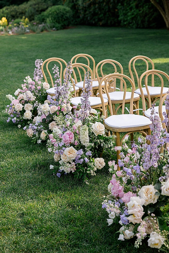 Ceremony aisle decor with a floral lined aisle of white and blush roses, pink peonies, and lavender delphinium beside bentwood chairs on grass