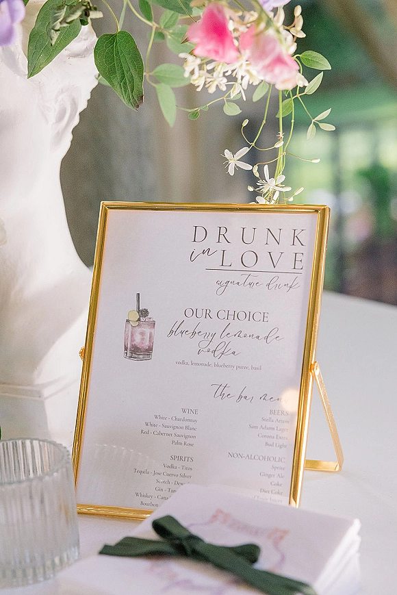 Wedding bar sign in a gold frame with a bar menu card and cocktail illustration on a white tablecloth in soft window light