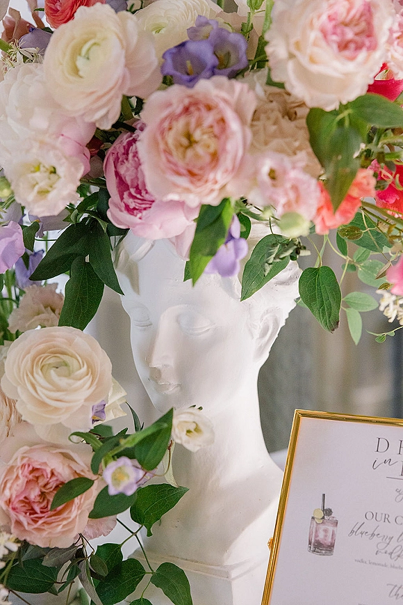 Wedding welcome sign in a gold frame with calligraphy, surrounded by roses, peonies, and purple blooms on a white tabletop with a bust statue