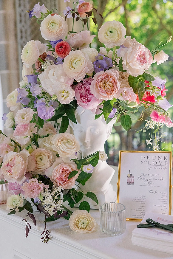 Wedding floral arrangement in a white urn with pastel blooms and greenery beside a gold-framed bar menu sign on a white tabletop by a window