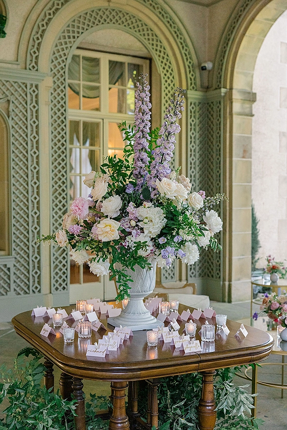 Wedding escort card table with escort card display, tall urn centerpiece, calligraphy place cards and votive candles by an arched doorway indoors