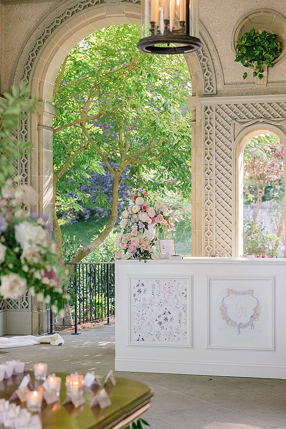 Wedding bar setup with a white wedding bar, floral arrangement, menu sign, and votive candles beneath a chandelier in an arched stone pavilion