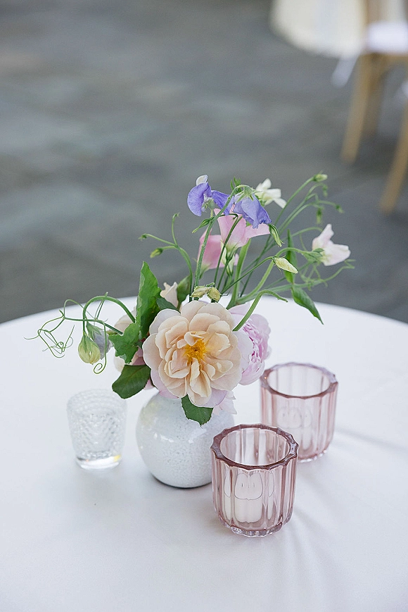 Wedding centerpiece with a bud vase of pastel sweet peas and garden roses, paired with pink glass votives on a white tablecloth at reception