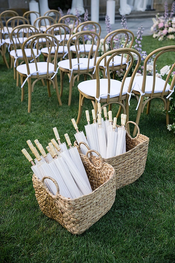Ceremony seating with outdoor wedding ceremony chairs, white cushions and ties, clear umbrellas in woven baskets on a grassy lawn aisle
