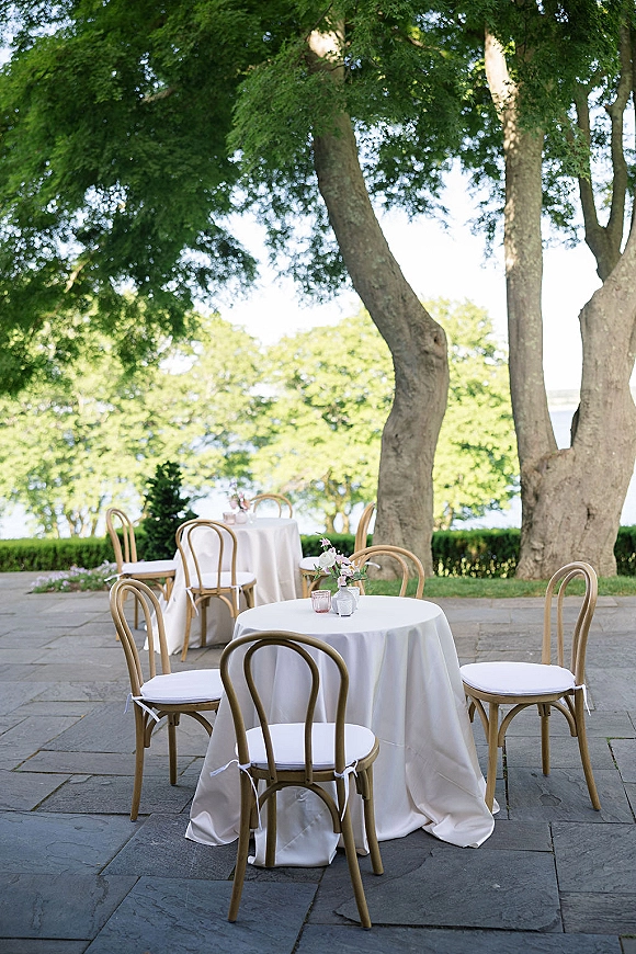 Outdoor reception tables with ivory tablecloths and bentwood chairs, set on a stone patio under trees with small floral centerpieces and votives