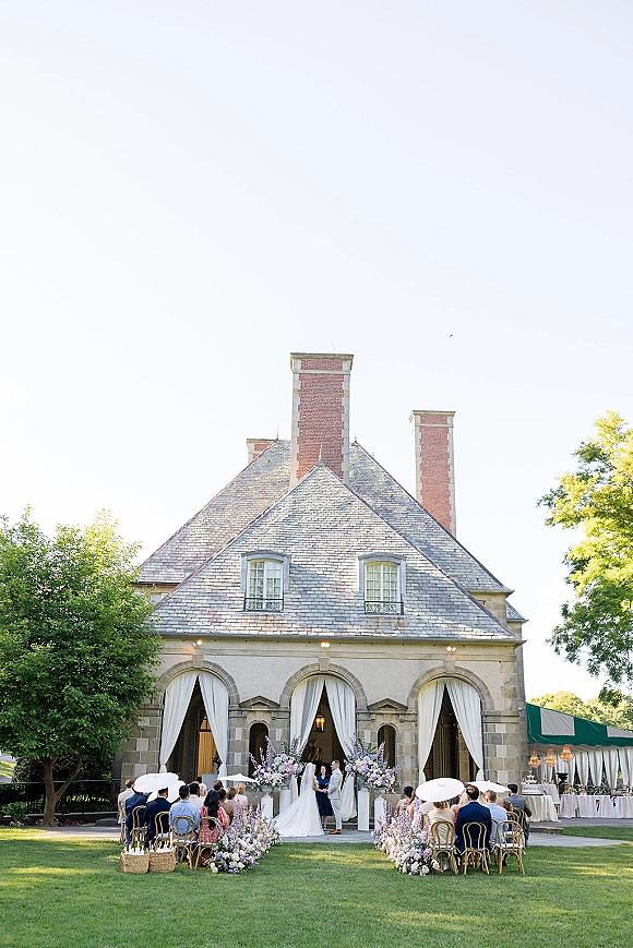 Outdoor wedding ceremony with floral lined aisle leading to a draped altar as bride in cathedral veil and guests with parasols sit by a stone estate facade