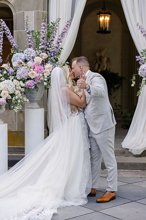 Wedding kiss portrait of bride and groom kiss on stone steps under an archway, her long veil trailing as he holds her waist