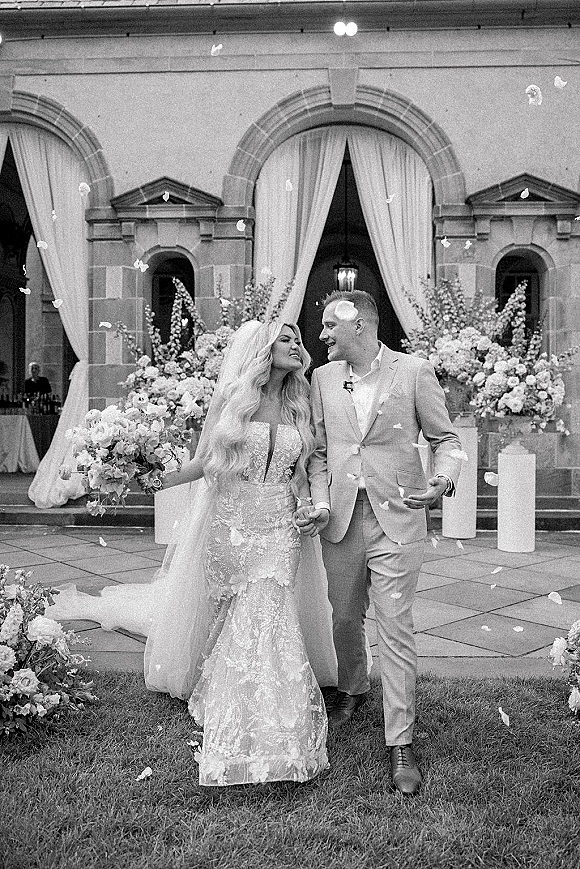 Wedding recessional as bride and groom walking hand in hand through a flower petal toss, her long veil and bouquet before stone arches